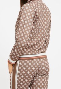 Chándal marrón con estampado y logos blancos, que incluye una chaqueta con cremallera, puños y cintura acanalados, y pantalones de tiro alto a juego.