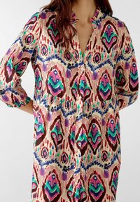 Camisa larga multicolor de tejido ligero que presenta un vibrante patrón ikat con acentos en púrpura, verde y azul, con botones al frente y cuello con volantes.