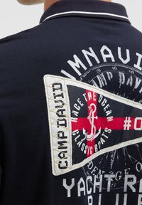 Navyblaues Poloshirt, Rückseite mit einem aufgestickten Patch, das ein rotes Kreuz, einen Anker und den Text "Camp David" sowie "Race the Ocean Classic Boats" zeigt.