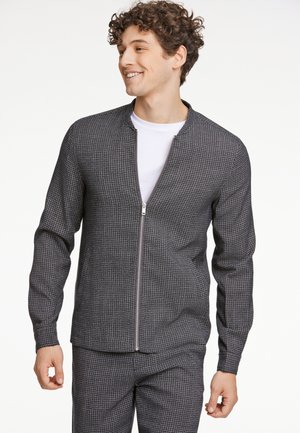 Harmaa vetoketjullinen takki, jossa on teksturoitu houndstooth-kuvio, pitkähihaisilla hihoilla ja pystykauluksella. Yhdistetty valkoiseen T-paitaan.