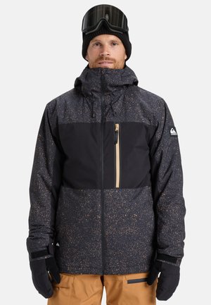 Man in een zwart en bruin gewatteerde winterjas, zwarte handschoenen, een zwarte gebreide muts met skibril, en tan sneeuwbroek tegen een witte achtergrond.