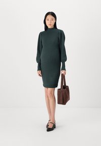 Robe en tricot de couleur vert foncé avec de longues manches bouffantes et un col haut, associée à un sac fourre-tout en suède marron, portée avec des chaussures à talons noires.