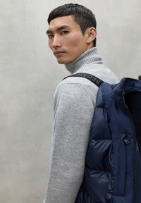 Suéter de cuello alto de punto gris combinado con un chaleco acolchado azul marino, que presenta una correa negra con el texto "BEC", texturas suaves y un diseño estructurado.