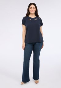 Námořnická modrá halenka s krátkým rukávem a výřezovými detaily kolem výstřihu, v kombinaci s tmavě modrými rozšířenými džíny a čirými sandály na podpatku.