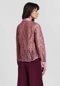 Pinker Spitzentop mit satinierter Kragen, das ein florales Muster aufweist. Lange Ärmel und ein leicht gewölbter Saum. Kombiniert mit dunklen Hosen.