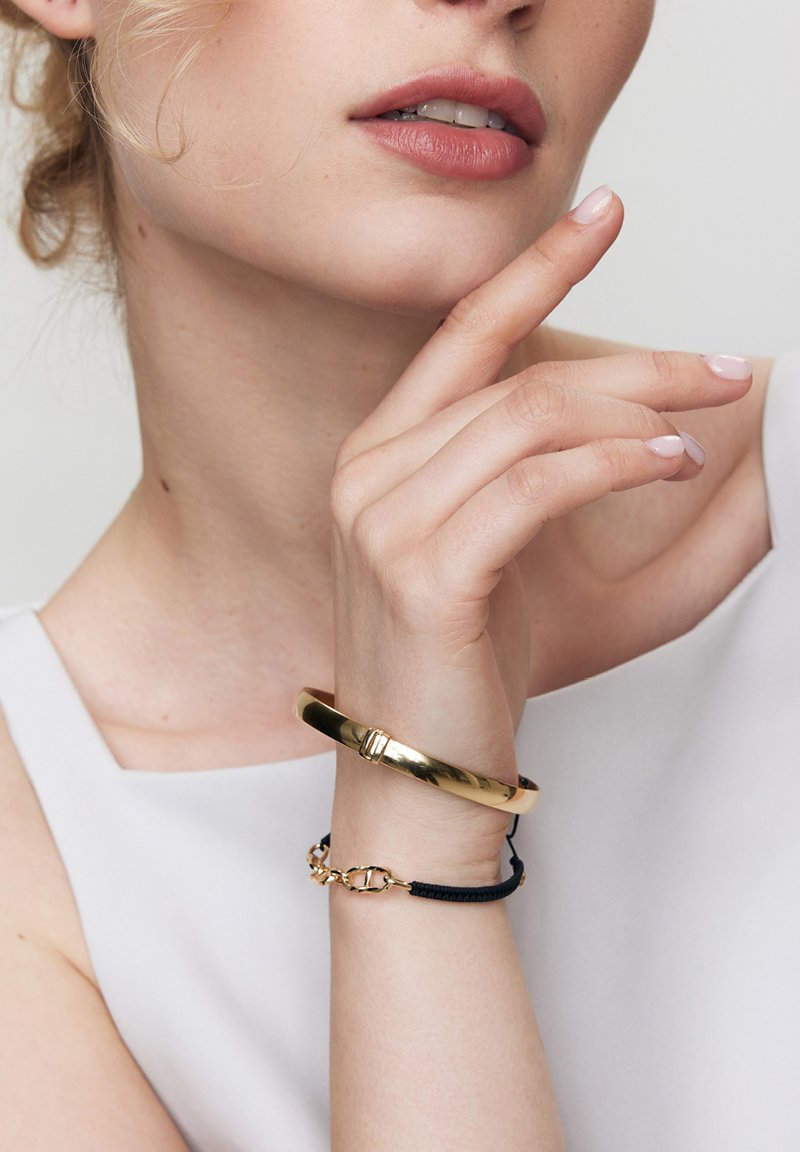 Złota bransoletka bangle o gładkim wykończeniu z małym akcentem. Obok niej noszona jest czarna bransoletka z cordu ze złotym wykończeniem.