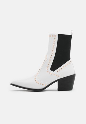 Bottines en simili cuir blanc avec des côtés élastiques noirs, bout pointu, ornées de clous en rose gold et avec un talon en bois.