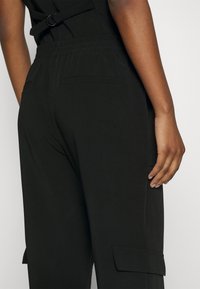 Un pantalon cargo noir présente une taille élastique, deux poches arrière et une texture lisse, avec des poches latérales pour plus de commodité.