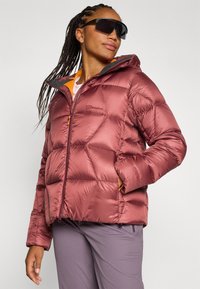 Mujer con trenzas lleva una chaqueta acolchada color burdeos y gafas de sol, posando con confianza frente a un fondo gris claro.