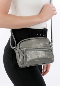 Metallic sidabrinė per petį nešama rankinė su užtrauktuko priekiniu kišeniu, minkštu paviršiumi, pūkuotu dekoru ir reguliuojama juosta, vilkima prie juodos aprangos.