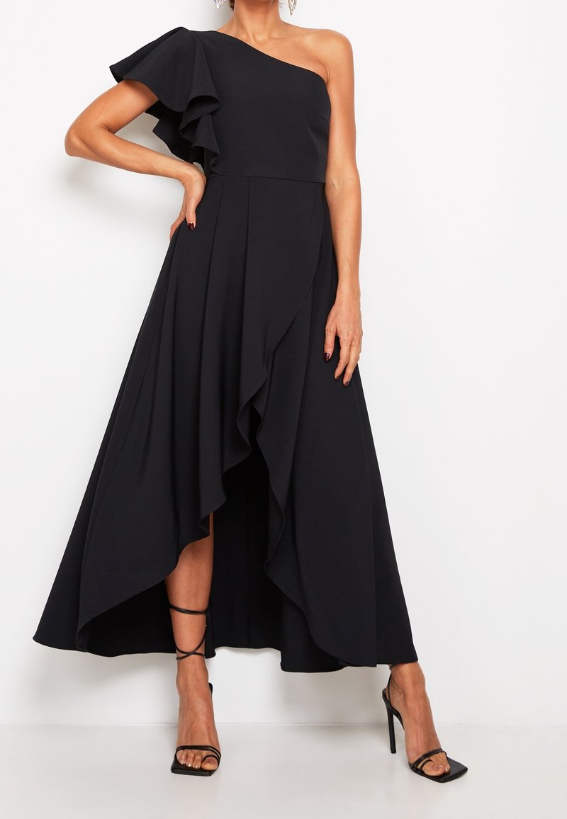Femme portant une robe noire asymétrique à une épaule avec un détail volanté sur l'épaule, associée à des sandales noires à talons hauts.
