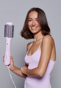 Brosse à cheveux ronde avec des poils noirs, un manche rose et un bouton d'alimentation. Le modèle la tient tout en portant un débardeur à côtes de couleur violet clair.