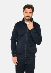 Felpa con zip di colore navy realizzata in morbido tessuto felpato, caratterizzata da un colletto alto, polsini a costine e un design minimalista senza motivi.