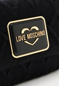 Melns teksturēts auduma soma ar zelta tonalitātes taisnstūrveida logotipa plati, uz kuras ir rakstīts "LOVE MOSCHINO" un sirds grafika.