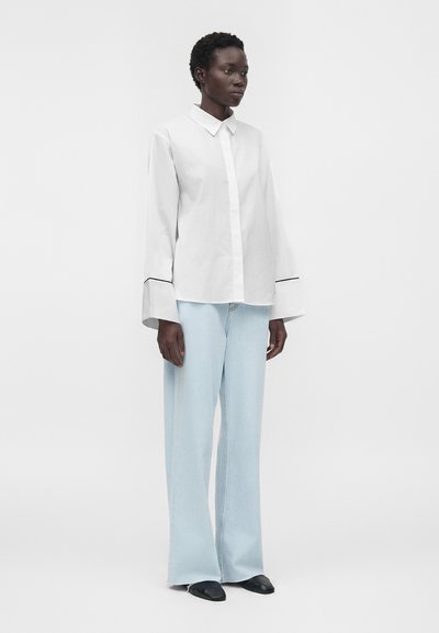 Personne noire portant une chemise blanche à manches longues avec de larges poignets et un jean bleu clair à jambes larges, debout devant un fond clair uni.