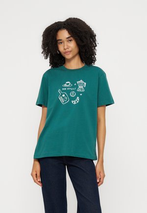 Femme portant un t-shirt bleu sarcelle avec des illustrations blanches sur le thème du petit déjeuner et le texte "Bon Appetit", associé à un jean foncé sur fond blanc.