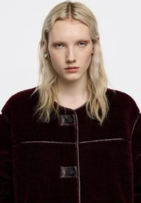 Chaqueta de forro polar burdeos con cuello redondo, que presenta un cierre frontal con botones y detalles en cuero marrón en contraste. Superficie texturizada con costuras visibles.