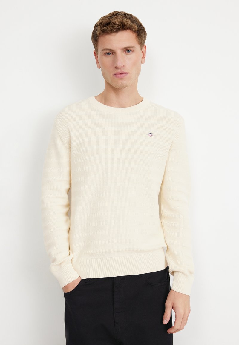 Jasny kremowy sweter z poziomą struktura prążków, okrągłym dekoltem i dopasowanymi mankietami. Małe haftowane logo na piersi. Czarne spodnie.