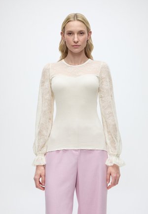 Kobieta o długich blond włosach nosi białą bluzkę z przezroczystymi rękawami z kwiatowej koronki oraz jasnoróżowe spodnie o szerokich nogawkach, stoi na jednolitym tle.