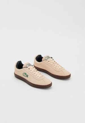 Par beige lave sneakers med sort indre foring, brune såler, snørebånd og en grøn krokodillelogo på siden.