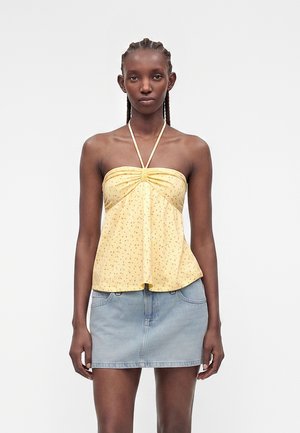 Femeie cu părul împletit purtând un top galben halter cu imprimeu floral mic și o mini-fustă din denim albastru deschis, stând pe un fundal simplu.