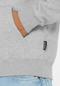 Gray sweatshirt with ribbed cuffs and hem, side pocket detail, and a black brand label on the lower side. Soft texture, casual design.
