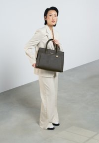 Sac à main structuré marron foncé avec une surface texturée et des accents en doré, tenu par une personne en costume clair.