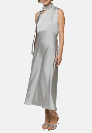 Donna che indossa un vestito midi senza maniche in satin argento con collo alto annodato e sandali argentati con tacco alto a listini, sorridente.