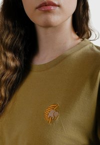 Camiseta de algodón verde oliva con un pequeño logo bordado en naranja y beige en el pecho. Cuello redondo con una textura suave.