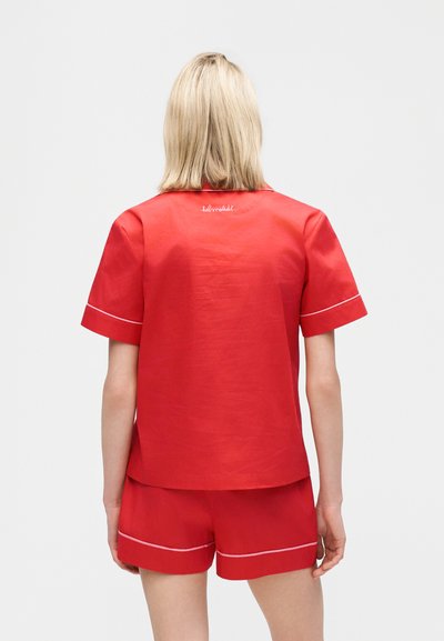 Blonde Person trägt rotes kurzärmeliges Oberteil und passende Shorts mit weißem Besatz, von hinten vor einem schlichten Hintergrund gezeigt.