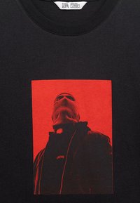 Čierne tričko s červeným štvorcovým grafickým motívom osoby v lyžiarskej maske a tmavom oblečení, ktorá sa pozerá nahor, s textom "UNFAIR" na hrudi.