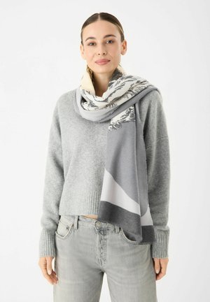 Écharpe en tricot grise avec un motif mixte de gris clair, gris foncé et crème. Présente une texture douce et un bord frangé. Portée drapée sur un pull.