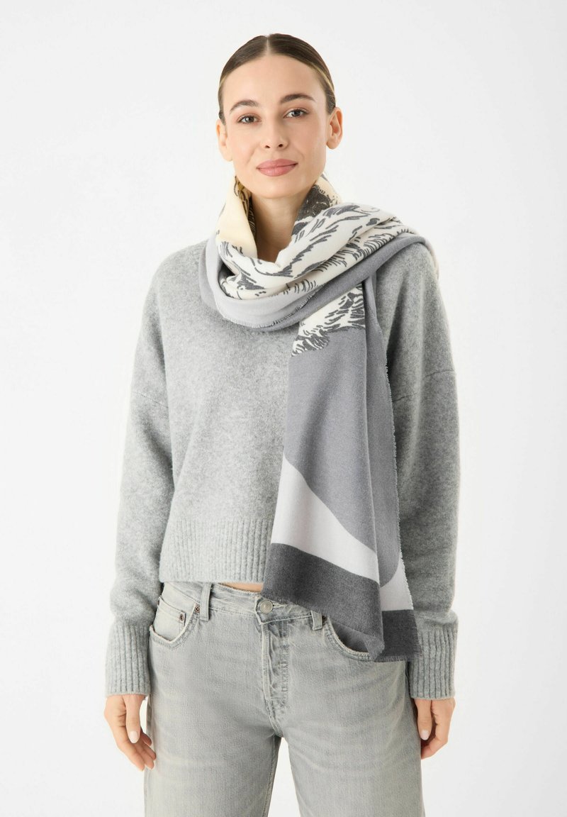 Écharpe en tricot grise avec un motif mixte de gris clair, gris foncé et crème. Présente une texture douce et un bord frangé. Portée drapée sur un pull.
