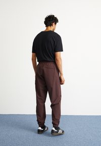 Melns T-krekls, brūni joggeri ar elastīgu jostasvietu un aizmugurējo kabatu, kā arī melnas kedas ar baltām svītrām. Vienkārša fona un zila grīda.