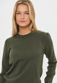 Suéter de punto en verde oscuro, con un cuello redondo acanalado y mangas largas con sutiles hombros abullonados; textura suave y diseño ajustado.