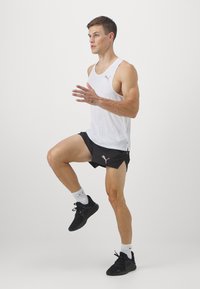 Outfit sportivo da uomo con top bianco senza maniche e pantaloni corti neri con spacchi laterali. Scarpe da corsa nere e calze bianche completano il look.