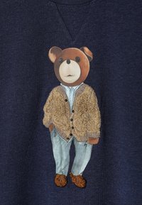 Tumši zils sporta krekls ar lāča ilustr�āciju, kas ģērbies bēša kardigānā, gaiši zilā kreklā un mintzaļās biksēs ar brūnām kurpēm.