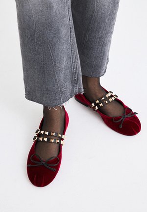 Bailarinas de terciopelo rojo con un lazo negro, adornadas con correas con tachuelas doradas. Llevadas con jeans de pierna ancha gris desgastados y medias negras.