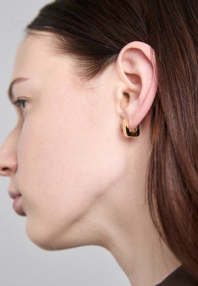 Goldfarbene quadratische Ohrringe mit hochglänzender Oberfläche, befestigt am Ohrläppchen, die das geometrische Design und die elegante Textur hervorheben.