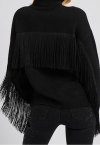 Femme portant un pull noir à franges et un jean noir avec des détails cloutés, vue de dos sur un fond uni.