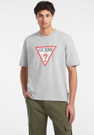 Grå bomullströja med en röd triangellogo som har "GUESS" och ett frågetecken i svart text. Korta ärmar och avslappnad passform.