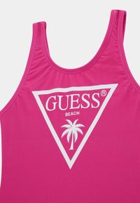 Costume da bagno fucsia con scollatura rotonda e spalline larghe. Mostra un logo triangolare bianco con la scritta "GUESS BEACH" e un grafica di una palma.