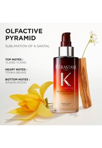 Il siero Kérastase 8H Magic Night in una bottiglia di vetro sfumata presenta una tonalità dal rosso scuro al chiaro, con un erogatore e accompagnato da un fiore giallo e un pezzo di legno.