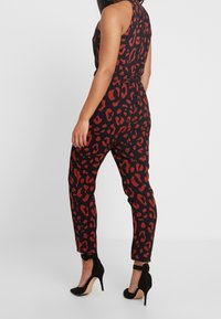Mono negro con estampado de leopardo rojo, diseño sin mangas, cintura ajustada, piernas ceñidas, combinado con zapatos de tacón negros.