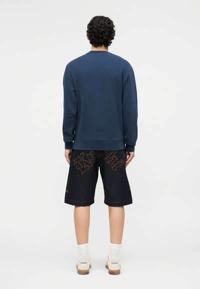 Άνδρας με σγουρά σκούρα μαλλιά, που φοράει μπλε μακρυμάνικο πουκάμισο, μαύρα σορτς μέχρι το γόνατο με πορτοκαλί ραφές, λευκές κάλτσες και αθλητικά παπούτσια, γυρισμένος με την πλάτη.