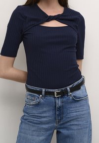 Námorně modrý žebrovaný top s krátkým rukávem a zkrouceným předním dílem, odhalující malý výřez pod výstřihem. V kombinaci se světle modrými džíny a černým opaskem.