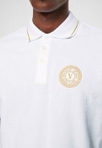 Polo blanc avec un petit logo doré brodé sur la poitrine gauche, doté d'un col classique et d'une patte de boutonnière à trois boutons.