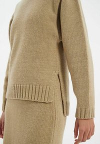 Pull en tricot beige avec un ourlet côtelé et des fentes sur les côtés. Présente un tissu texturé et des manches longues, associé à une jupe assortie.