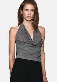 Grijze haltertop gemaakt van lichte stof, met een gedrapeerd ontwerp, subtiele versieringen en een getailleerde pasvorm. Gecombineerd met zwarte broeken.