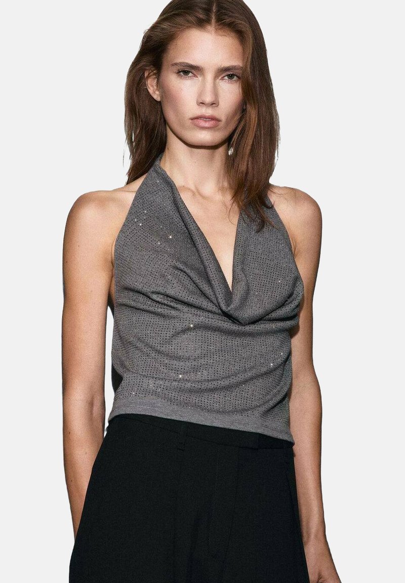 Grijze haltertop gemaakt van lichte stof, met een gedrapeerd ontwerp, subtiele versieringen en een getailleerde pasvorm. Gecombineerd met zwarte broeken.